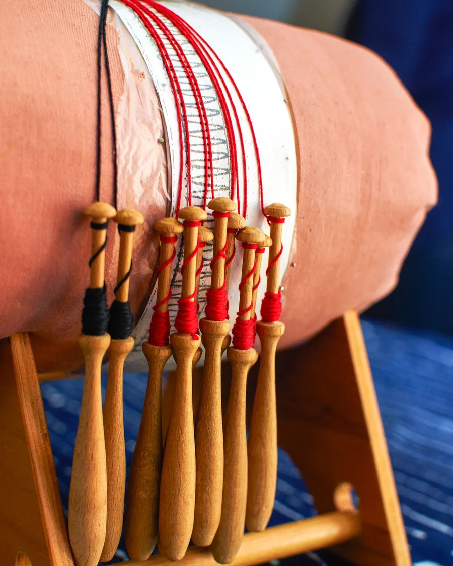 Bolillos de madera con hilo rojo y negro devanado, colgando de un mundillo cilíndrico con el picado sujeto